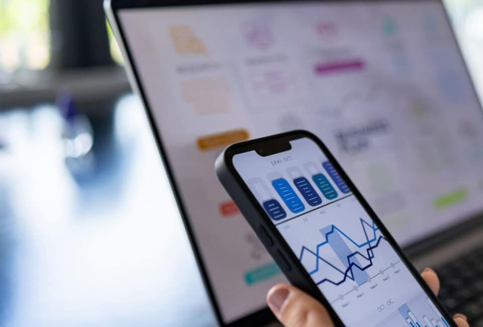 A person using a laptop and smartphone to analyze financial data and charts.